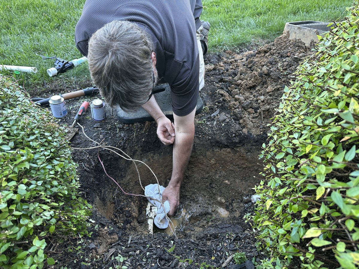 Josh kneeling beside an excavated valve box, cleaning the exposed pipe during valve replacement